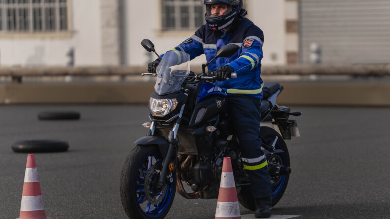Un motard de la gendarmerie sur sa moto en attente entre deux plots orange et blancs, derrière une ligne jaune, sur un plateau de moto