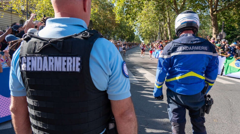 Un gendarme et un motard de la gendarmerie de dos devant une route ensoleill&eacute;e o&ugrave; courent des personnes, avec de part en d'autre la foule