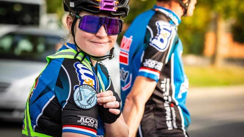 Agrandir l'image Une jeune femme cycliste portant un casque, des lunettes violettes et ue combinaison de cycliste nous regarde, souriante, la main gauche désignant l'écusson sur lequel est écrit REALISE 2023 apposé sur son épaule droite