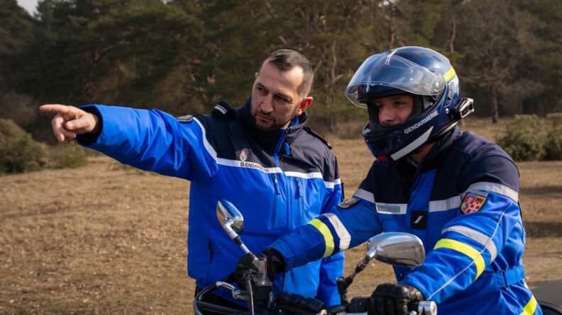 Un instructeur gendarme pointe de la main droite quelque chose au loin, à un motard de la gendarmerie, casqué, qui regarde. EN arrière-plan, un terrain sauvage et une forêt de résineux