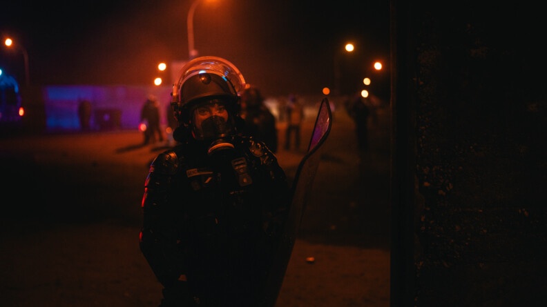 Une femme gendarme, portant une tenue noire, un casque et un masque, se tient debout, face à l'objectif. La scène se déroule de nuit.