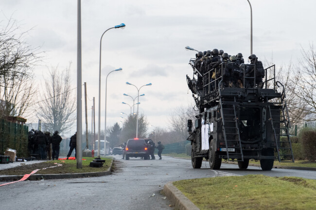 Progression du véhicule SHERPA transportant des équipiers du GIGN. Devant eux, deux équipiers sont abrités derrière un autre véhicule noir. A gauche, contre la haie, une colonne d'assaut.