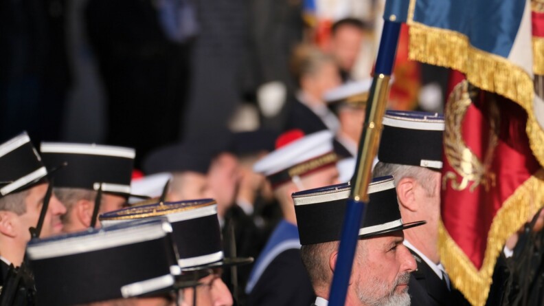 Des militaires se tiennent côte à côte. Ils sont porstionnés de biais par rapport aux photographe. Ils sont en gros plan. On aperçoit que leur tête et leurs épaules. Ils sont vêtus d'une tenue sombre et protent un képi noir gansé de blanc. L'un d'eux porte des gants blancs. Il tient dans ses mains le mât d'un drapeau.