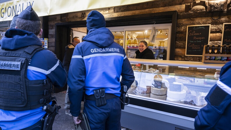 Deux gendarmes de dos échanges avec une commercante placée de l'autre côté d'une vitrine de fromage et devant son magasin en bois et baie vitrée, souriante