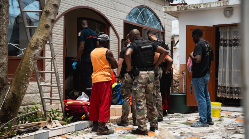 Trois gendarmes en treillis et hauts noirs avec des civils et des pompiers à l'entrée d'une maison