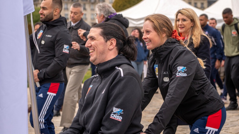 Marjorie Delassus, avec le porteur du drapeau des Jeux Paralympiques, en fauteuil roulant, ils sont souriants et se dirigent vers l'h&ocirc;tel national des invalides
