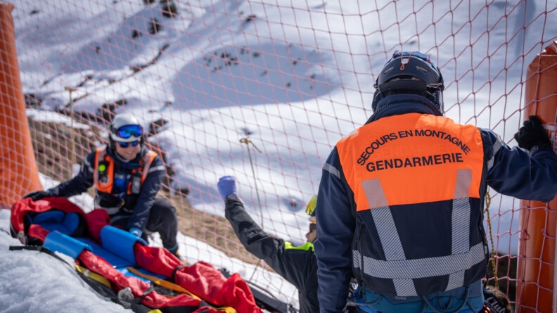 Trois gendarmes secouristes préparent le brancard pour un installer une victime. On voit un gendarme de dos avec noté sur sa chassuble "Secours en montagne Gendarmerie".