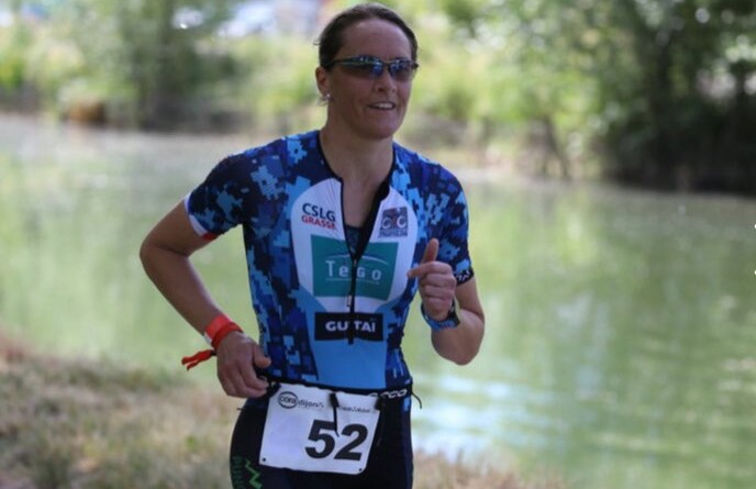 Lunettes de soleil sur le nez, l'adjudant Maud, vêtue d'un cycliste bleu foncé et d'un tee-shirt dans les tons de bleu affichant le dossard 52, en train de courir au bord d'une rivière bordée d'arbres..