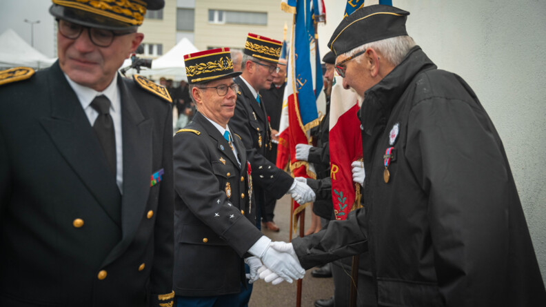 Le major g&eacute;n&eacute;ral de la gendarmerie nationale serre la main d'un homme se tenant de profil, portant une m&eacute;daille sur son manteau noir
