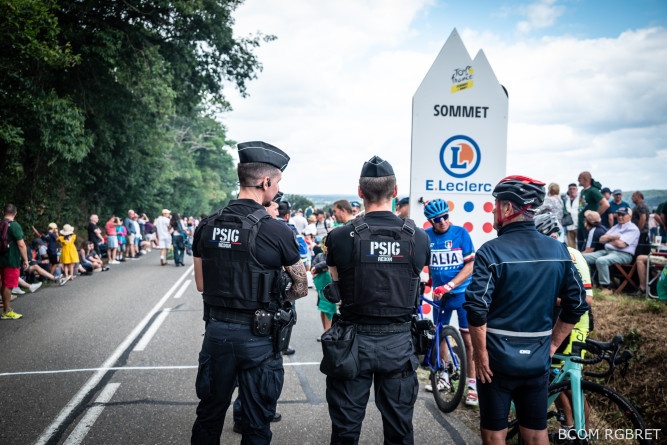 Agrandir l'image Deux gendarmes d'un Peloton de surveillance et d'intervention de gendarmerie (PSIG), vus de dos, sont au bord de la route. Ils échangent avec un cycliste amateur, casque sur la tête et vélo à la main, vu de dos. Au bord de la route, de part et d'autre de la chaussée, sont massés de nombreux spectateurs, debout et assis.
