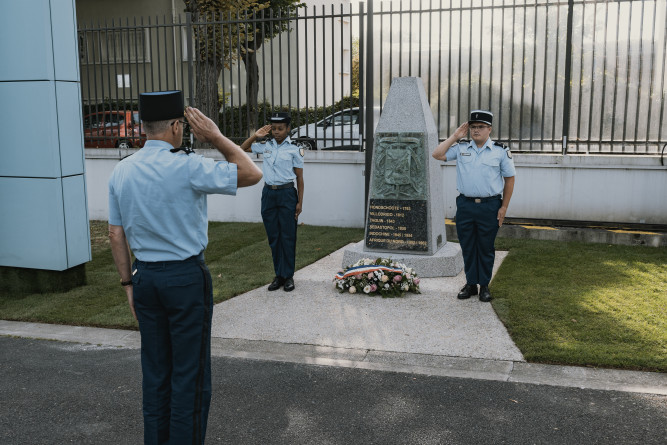 Trois gendarmes se recueillent en effectuant le salut militaire devant une gerbe de fleurs posée au sol, devant un monument mémoriel. La scène se déroule en journée, dans une cour.