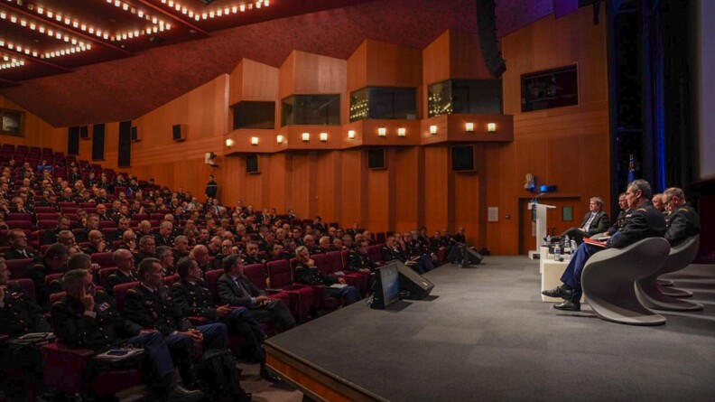 Sur une sc&egrave;ne, &agrave; droite, le directeur de la gendarmerie, le major g&eacute;n&eacute;ral et les diff&eacute;rents directeurs. A gauche, dans une grande salle en gradin, des officiers de gendarmerie en vareuse, install&eacute;s dans des fauteuils rouges.