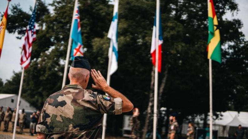 Agrandir l'image Un militaire, debout, de dos, en tenue kakie, effectue le salut militaire, devant les drapeaux de plusieurs pays.