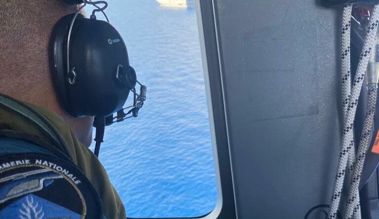 Vue sur la mer et le porte hélicoptères Mistral depuis le hublot d'un hélicoptère de la gendarmerie nationale. Au premier plan un militaire de l'équipage.