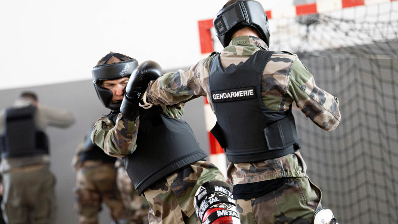 Combat de boxe dans un gymnase entre deux élèves gendarmes en treillis kaki, avec gilet pare-balles, casque de boxe, gants et jambières.
