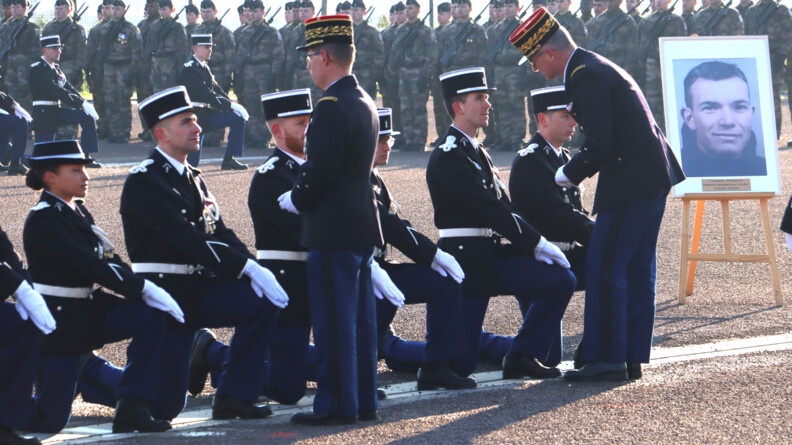 En arrière plan, des élèves-gendarmes en treillis, famas en main, au garde-à-vous.  Devant eux, une première rangée d'élèves-gendarmes en vareuse, agenouillés. Au premier plan, une rangée de sept élèves-gendarmes, en vareuse, agenouillés, auxquels le major général et le commandant de région, de dos, remettent successivement les galons. A droite, un portrait, sur chevalet, du parrain de promotion.
