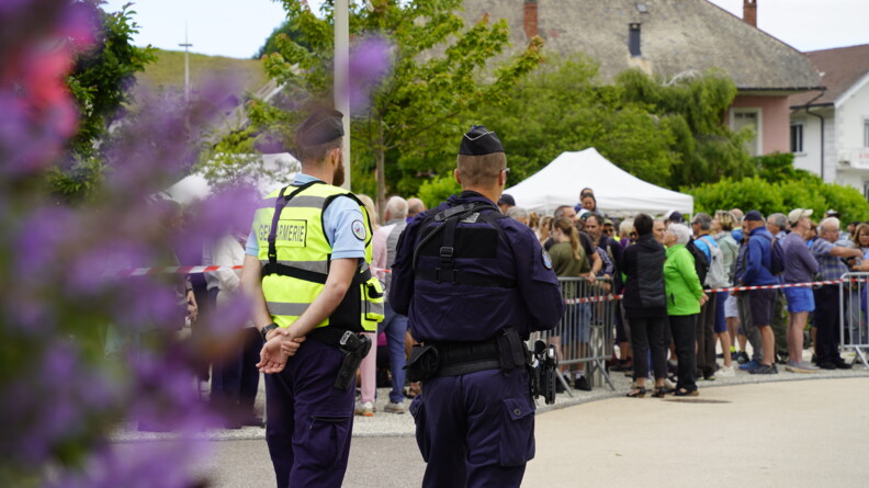 De dos, une gendarme de brigade et u nautre de PSIG attendent sur une route, de nombreuses personnes en sécurité derrière une barrière