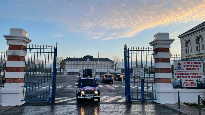 Une 4L bleue marine franchit les grilles d'enceinte d'un grand b&acirc;timent beige au toit sombre. Elle quitte le lieu, et pr&ecirc;te &agrave; s'engager sur la route. Le ciel est bleu clair, constell&eacute; de petits nuages.
