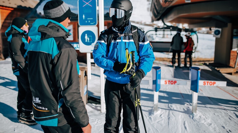 Un gendarme en tenue de ski, de face, tenant deux bâtons de ski dans ses mains, échange avec deux hommes en tenue de ski noir et bleu à l'entrée de remontées mécaniques.