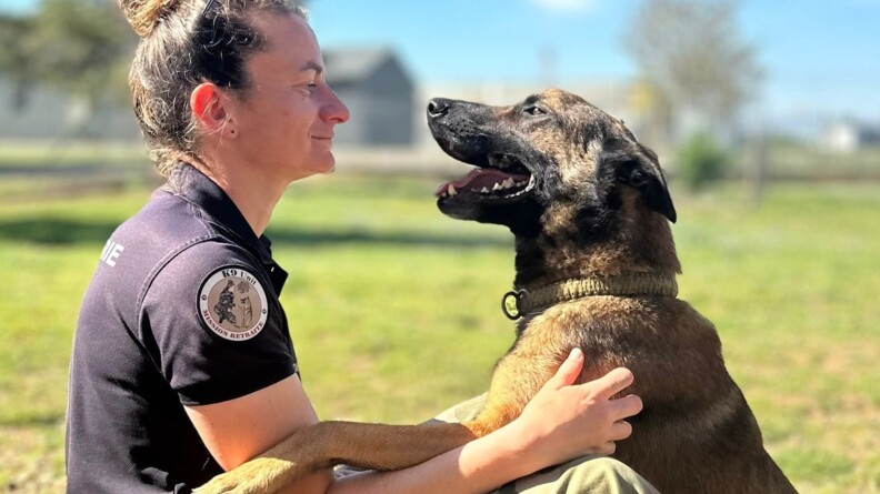 Photo d'une femme gendarme, assise sur la pelouse, de biais par rapport &agrave; l'objectif, face &agrave; son malinois, qu'elle tient au niveau des &eacute;paules