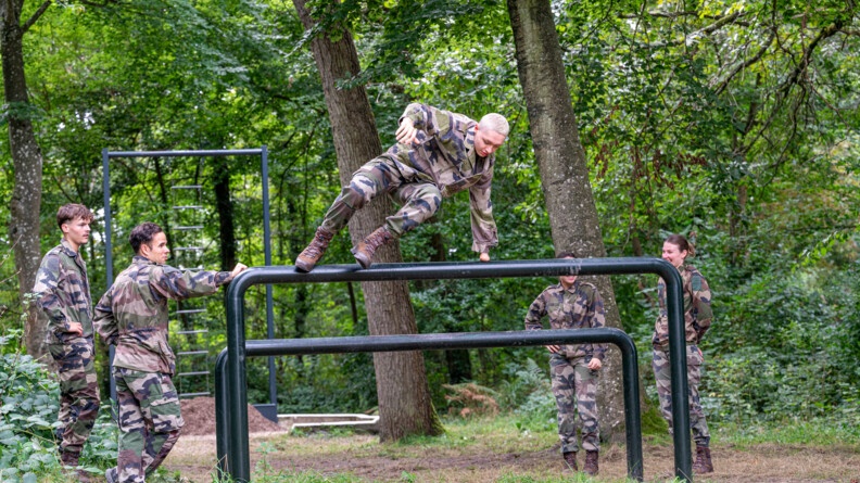 Quatre personnes en treillis, une cinquième, a qui il manque une main, passant par-dessus un obstacle, dans la forêt