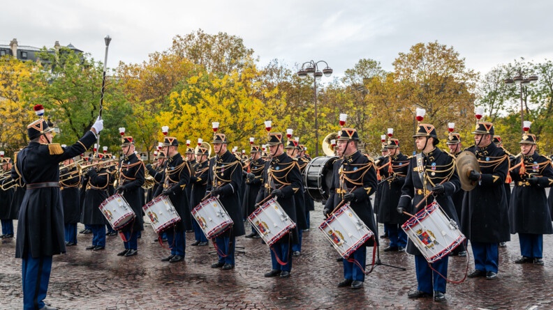 Devant des arbres dont les feuilles sont de la couleur de l'automne, sur un sol pavé, se tient un orchestre de la Garde républicaine. Certains ont un tambour, et d'autres des trompettes. Un chef d'orchestre qui se tient debout devant eux dirige l'ensemble. Au-dessus d'eux, le ciel est gris.