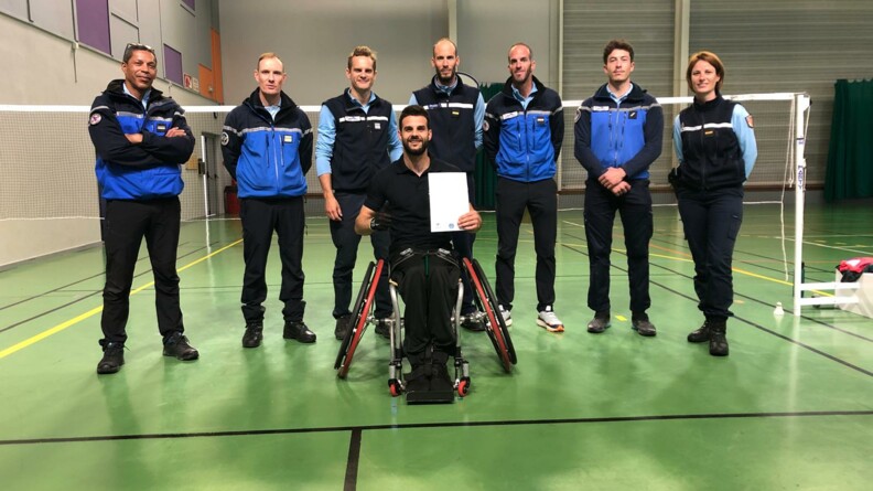 Thomas Jakobs et les militaires du PGHM se tenant devant le filet de badminton après l'entrainement. Thomas tient la convention dans ses mains.