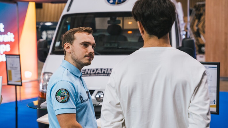 Un gendarme vêtu d'un polo bleu ciel discute un homme brun, vêtu d'un t-shirt à manches longues de couleur blanche, se tenant également debout, de dos. On aperçoit en arrière plan une camionnette blanche de la gendarmerie. La scène se déroule au salon de l'Auto 2024, à Paris.