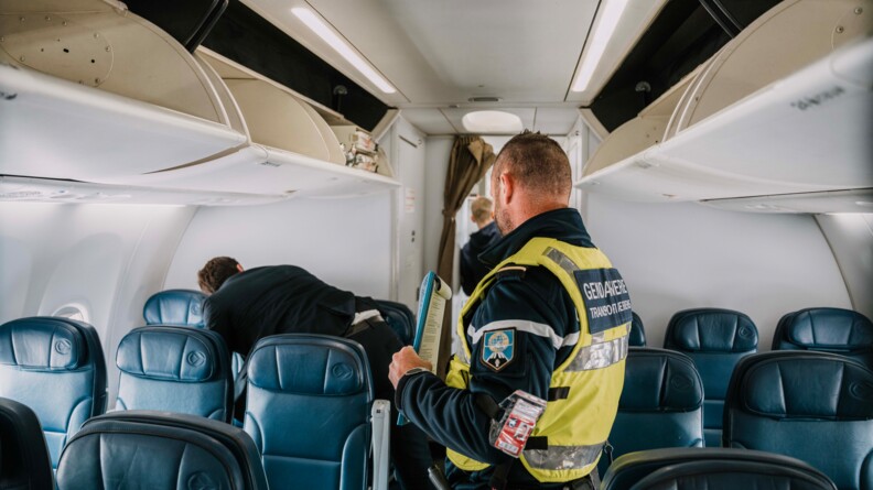 Un gendarme se tenant de baiais, revêtant un gilet chasuble jaune sur son uniforme, avec la mention dans le dos "Gendarmerie Transports aériens", tient dans sa main gauche un document. Il se trouve dans l'allée centrale d'un avion de ligne, dont les sièges sont vides. Il observe un steward effectuant un contrôle au niveau des sièges situés tout au fond de l'appareil.