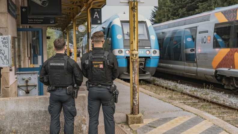 La sc&egrave;ne repr&eacute;sente deux gendarmes, debout, de dos, sur un quai de gare, alors q'un train de couleur bleue et blanche entre en gare.