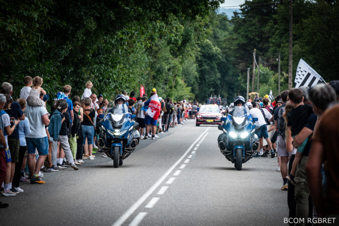Agrandir l'image Deux motocyclistes de la gendarmerie ouvrent la route d’une course cycliste féminine, suivis de près par un véhicule de la course et un peloton de coureuses. Les motos bleues sont équipées de gyrophares allumés. De nombreux spectateurs bordent la route des deux côtés, applaudissant et filmant les cyclistes. L’ambiance est festive et animée.