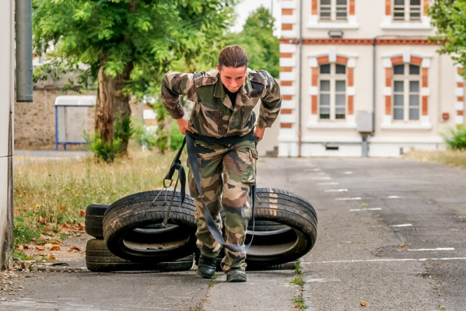 Agrandir l'image Un jeune militaire en tenue camouflage réalise un exercice de tractions de pneus, arrimés à sa taille par une large corde noire, sr un chemin en bitume. Derrière lui, en arrière plan, apparait un bâtiment en béton beige.