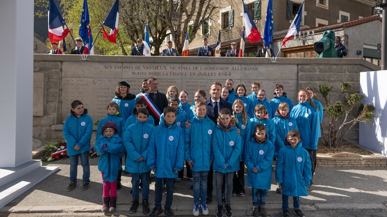 Agrandir l'image Le président de la république, Emmanuel Macron, apparait de face, debout, entouré d'enfants vêtus du même blouson bleu, devant un monument aux morts, sur lequel est inscrit "Aux enfants de Vassieux victimes de l'agression allemande morts pour la France en juillet 1944