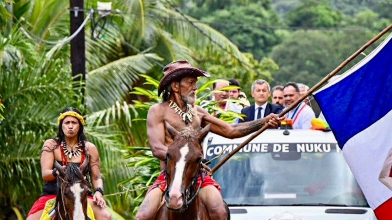 Le ministre de l'Int&eacute;rieur et des Outre-mer, G&eacute;rald Darmanin, debout dans une voiture dont le toit est ouvert, pr&eacute;c&eacute;d&eacute; par deux polyn&eacute;siens en tenue traditionnelle sur des chevaux. L'un porte un drapeau de la France.