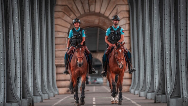 Deux gendarmes cavaliers évoluent sur leur monture, de grands chevaux de couleur brune arborant une tâche blanche sur le front, avançant sur une voie en bitume, encadrée par de grandes arcades métalliques.