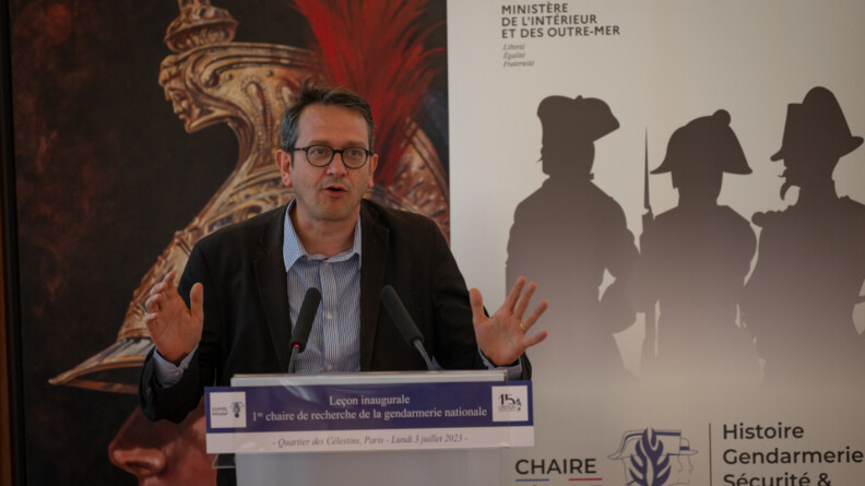 Le professeur Houte fait un discours au pupitre avec deux micros devant un panneau avec des silhouettes en ombre de gendarmes et une peinture d'un garde r&eacute;publicain.