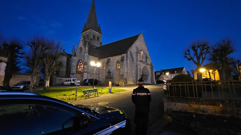 Au crépuscule, à la lumière des lampadaires. A gauche un véhicule gendarmerie, à sa droite au centre une gendarme de dos. A droite une grille. Un parvis d'église avec de lh'erbe, des arbres et des places de parking. En arrière plan légèrement sur la gauche ,une église en pierres grises