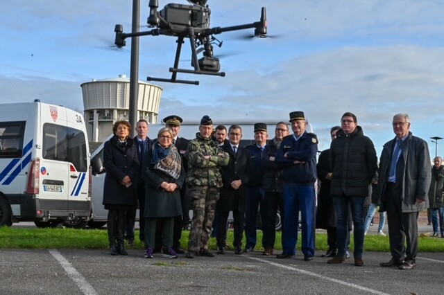 A l'ext&eacute;rieur, sur un sol b&eacute;tonn&eacute;, avec une pelouse derri&egrave;re eux, se tiennent debout face au photographe, des hommes et des femmes, militaires et civils. Tous regardent un drone qui vole devant eux. Derri&egrave;re eux, on aper&ccedil;oit une camionette blanche des forces de l'ordre, et le ciel bleu et gris.