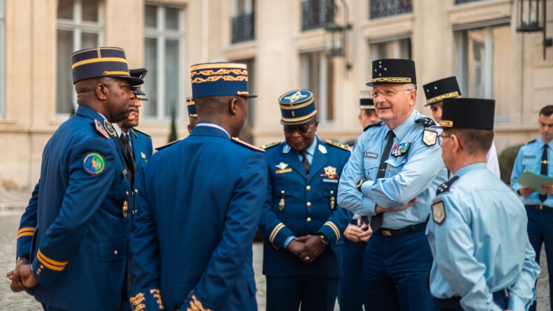 Agrandir l'image Dans une cour extérieure pavée se tiennent deux autorités de la gendarmerie nationale, entourées de gendarmes congolais. Derrière eux, se trouve un bâtiment formant un angle, dont on distingue les fenêtres