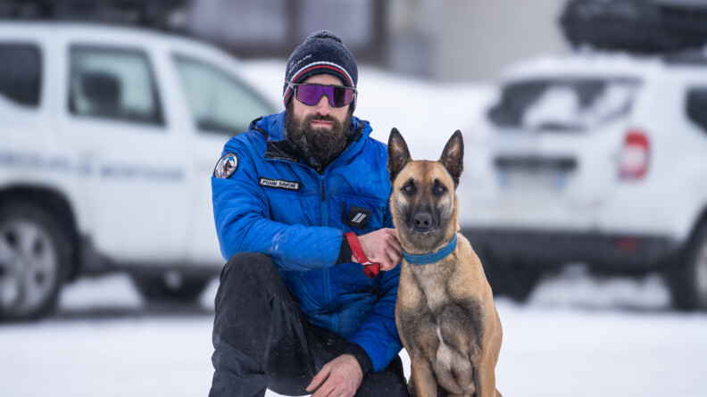Agrandir l'image Un gendarme se tient accroupi, un genou posé à terre, sur le sol enneigé. Il tient au collier de sa main droite un berger belge malinois, assis contre sa jambe gauche. L'homme porte une veste bleue portant sur la poitrine la mention PGHM Savoie et un écussion en haut de la manche droite sur lequel on aperçoit un chien. Derrière eux, on distingue des véhicules blancs floutés.