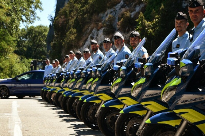 Agrandir l'image Un groupe de motocyclistes de la gendarmerie alignés côte à côte sur la route. En arrière plan apparaît un véhicule sérigraphié de la gendarmerie.