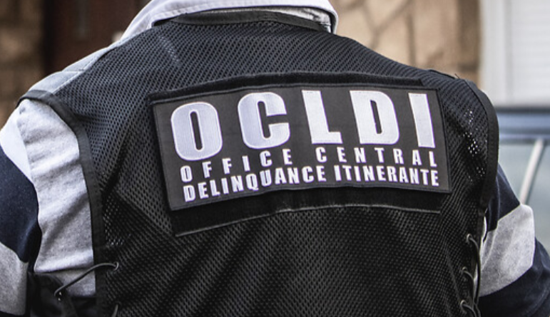 Vue de dos sur une chasuble noire siglée en blanc OCLDI - Office central délinquance itinérante, portée par un homme dont on voir les manches de polo grises et bleu marine