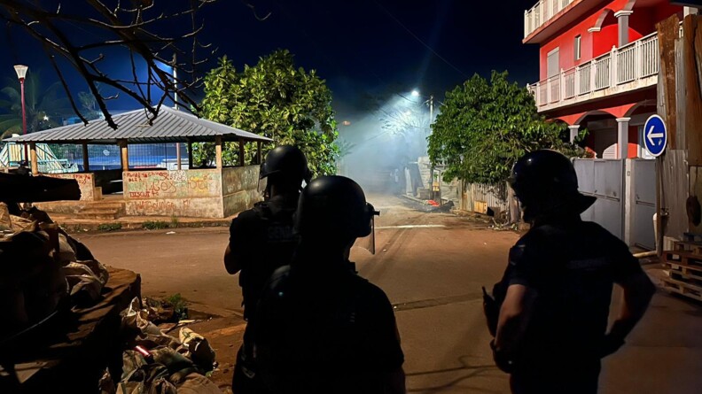 Trois gendarmes mobiles de dos, casqu&eacute;s, visi&egrave;res baiss&eacute;es, en patrouille de nuit dans une rue en terre battue. A leur gauche des immondices. Face &agrave; eux un b&acirc;timent en ruines recouvert de graffitis et plus loin des habitats en t&ocirc;le.