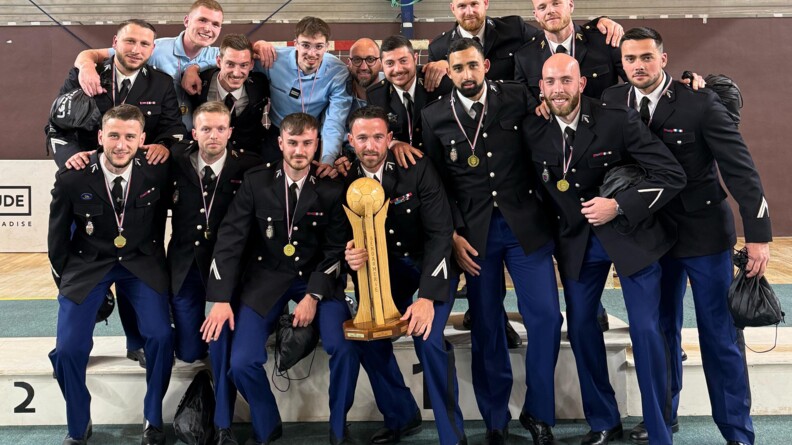 Agrandir l'image Quinze gendarmes, dont la majorité est en vareuse et les autres en tenue d'intervention, posent en groupe dans un gymnase, sur et devant un podium. Au milieu, l'un d'eux brandit un trophée. Tous ont une médaille autour du cou.