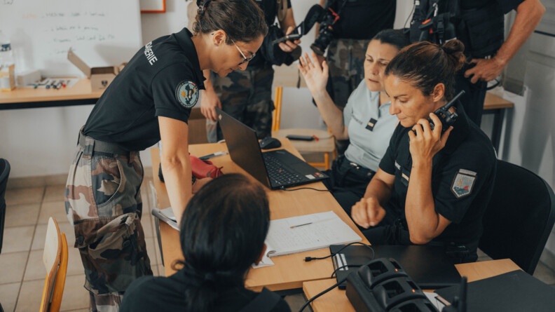 Plusieurs femmes gendarmes sont réunies autour d'une table sur laquelle sont posés un ordinateur et des cahiers. L'une d'elle a un talkie walkie à la main.
