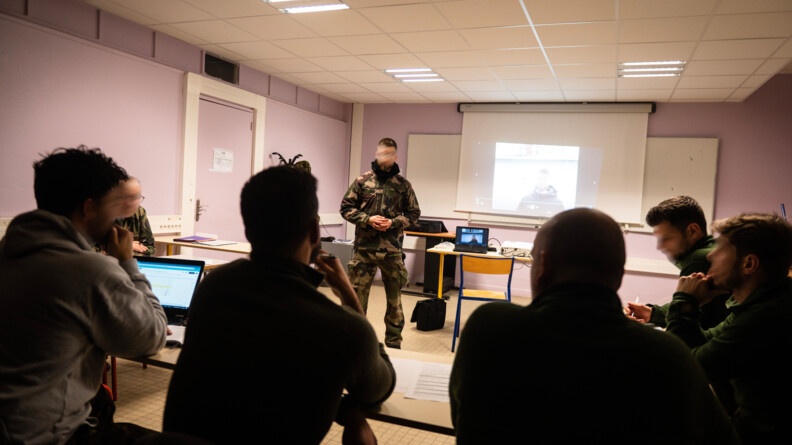 Les stagiaires sont cette fois en intérieur ou se déroule un autre volet de la formation. Dans une salle rose, face à un formateur, ils écoutent et observent les éléments projetés sur le tableau blanc par un rétroprojecteur.S