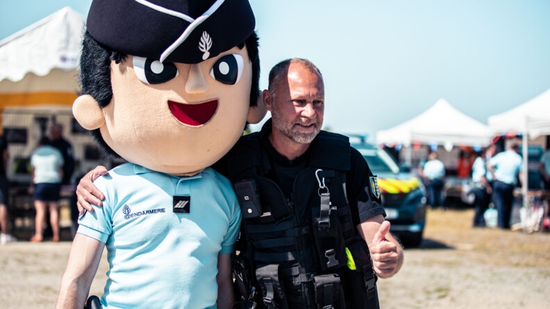 Un gendarme pose avec un autre gendarme qui porte la tête énorme de la mascotte, le gendarme Théo.