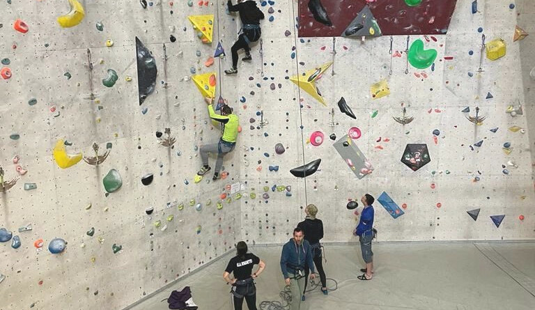 Quatre personnes se tiennent debout au pied d'un mur d'escalade, tandis que deux autres personnes montent le long du mur