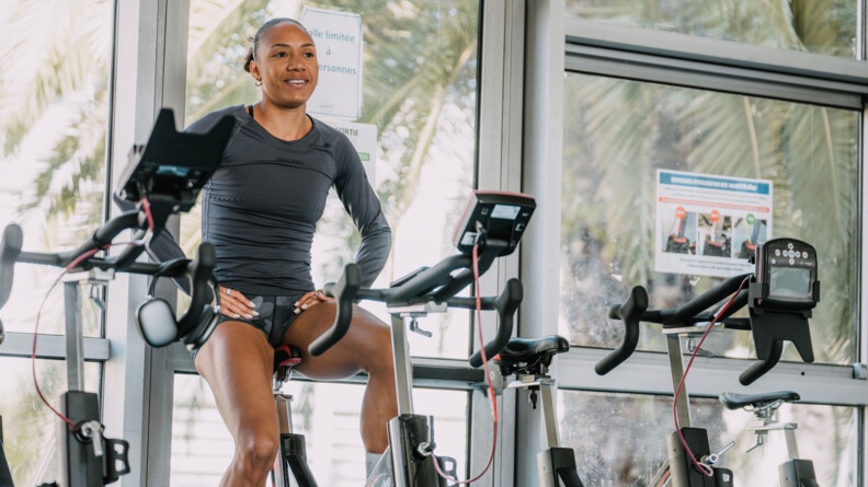 Une sportive vêtue d'une tenue grise, pédale, souriante, sur un vélo fixe. Ses mains sont posées sur le haut de ses cuisses.