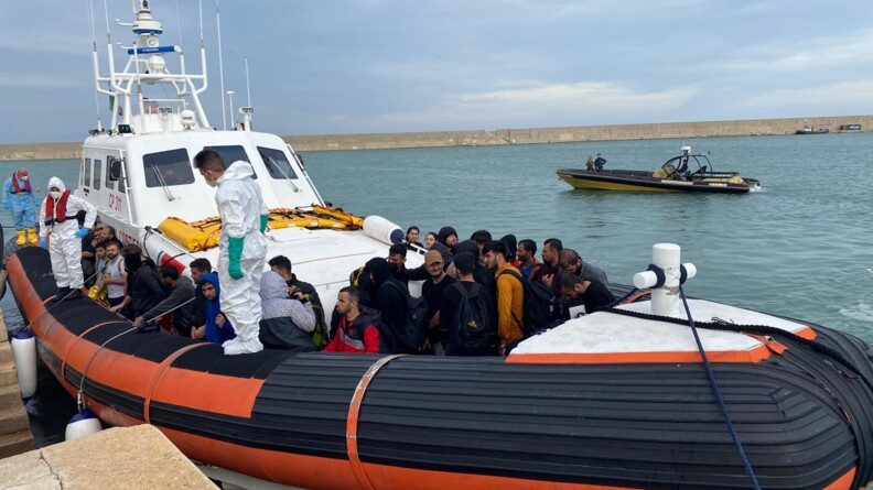 Agrandir l'image Un groupe de migrants rassemblés sur le pont d'un bateau à la coque banche et au bastingage orange et noir. Sur ce bastingage, côté quai, sont perchés deux hommes, en combinaisons blanches, gantés et masqués. En arrière-plan, un Zodiac jaune et noir, avec deux hommes à bord.